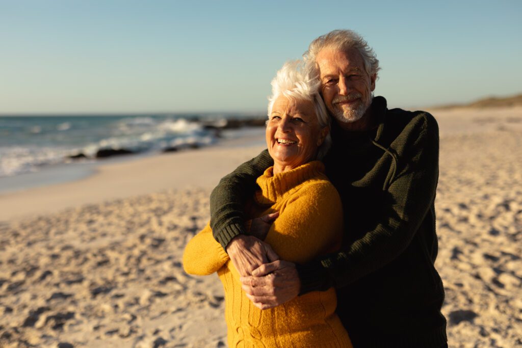 Front view of a senior Caucasian couple at the beach wearing sweaters, embracing and smiling to camera, with blue sky and sea in the background